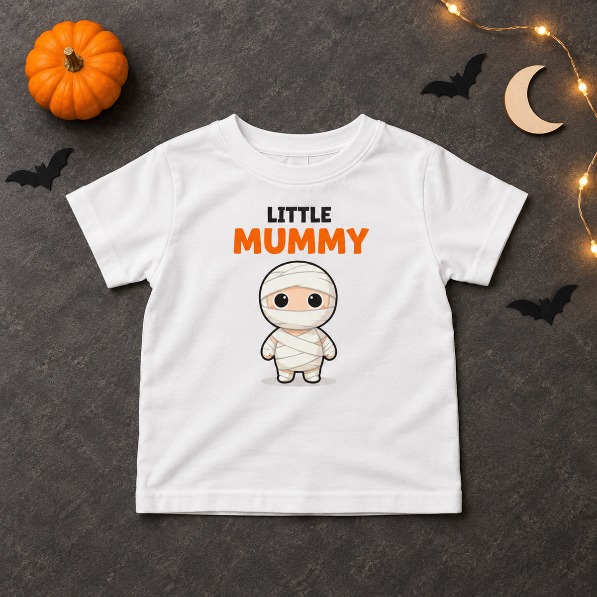 Little Mummy T-shirt flatlay with pumpkin, bats, moon, and warm Halloween lights